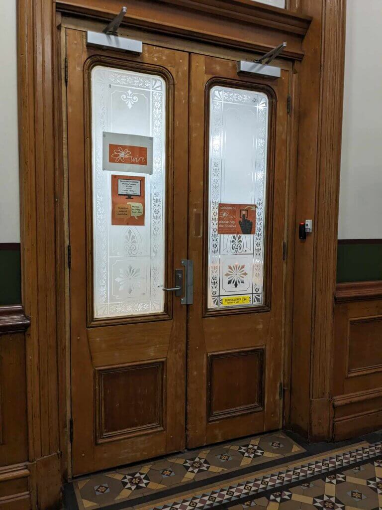 Photograph of Wire's front door. Two narrow wooden doors with a handle in the middle. There is a door bell to the right. The floor is Victorian era mosaic tiles.